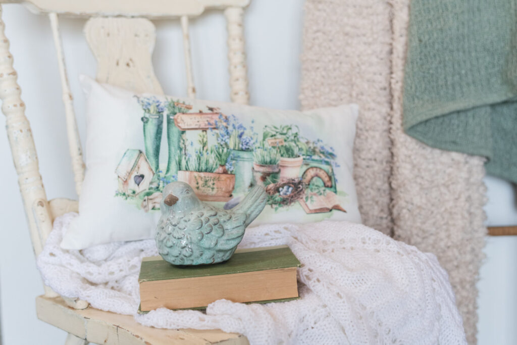 Soft, velvety, pillow sitting on a vintage chair in the corner of a bathroom draped with a blanket book and a cute bird