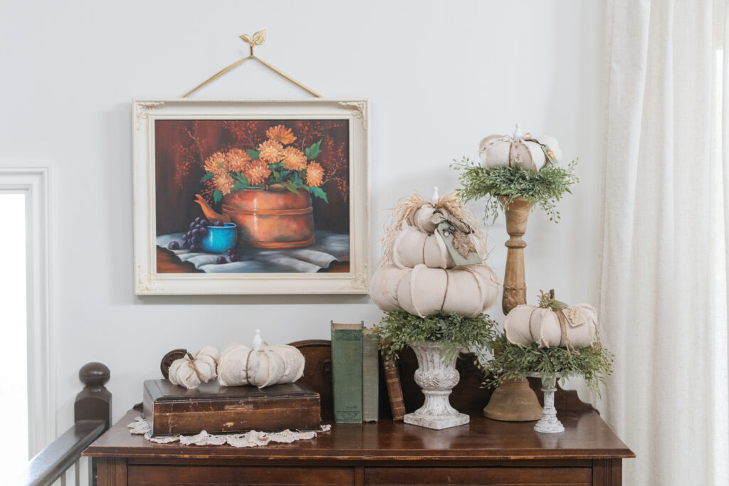 Drop cloth, pumpkin topiaries on candle sticks, urns and risers beside hand, painted wall art, and an old silverware case topped with more DIY pumpkins 