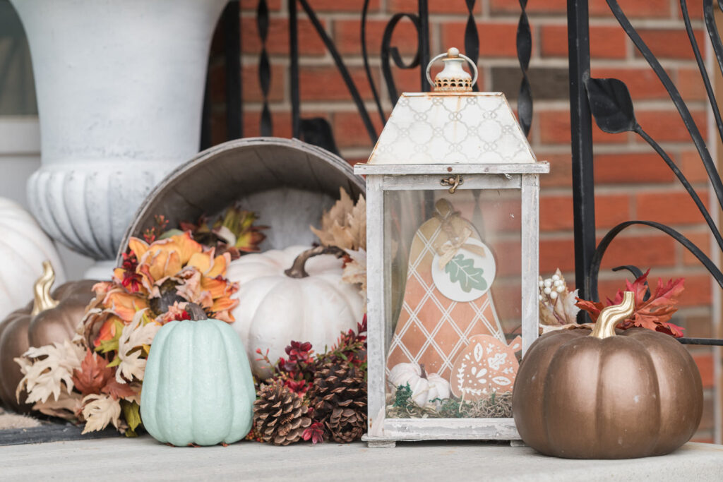 Vintage looking lantern with an apple basket, turned on its side, with pinecones and pumpkin, spilling out on the front step 