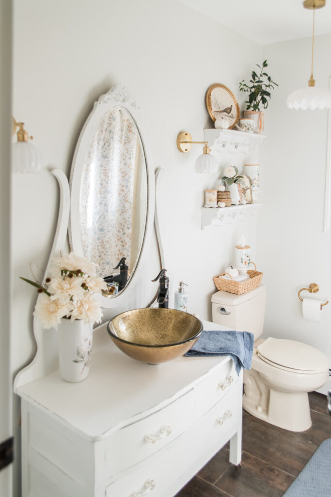 Bathroom makeover featuring updated brass lighting, a vintage dresser vanity with layered white paint, and autumn decor.