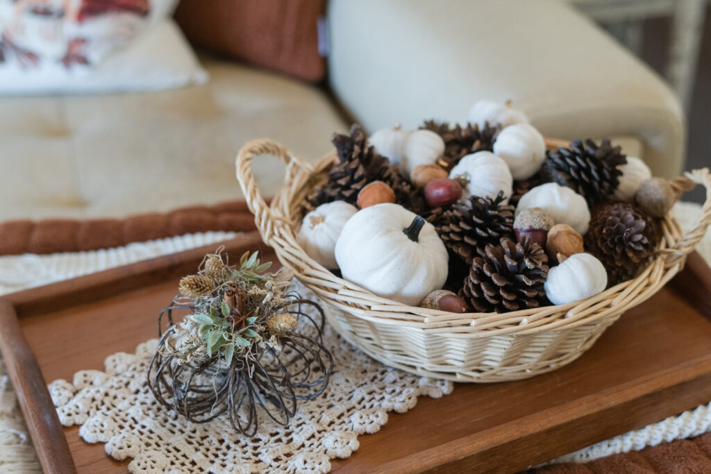 A wood tray with a basket of pumpkins and pinecones, with a jute pumpkin beside it, sitting on a doily for a cottage core feel 