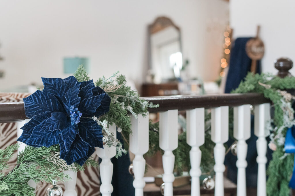 Soft-touch cedar garland with navy poinsettias and layered blue ribbons draped along staircase railing.