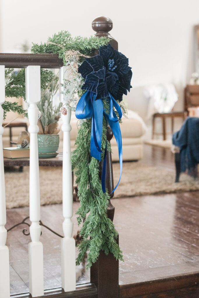 Both cedar garland drape down the end, post of a railing in an entryway with a blue velvet, poinsettia and velvet and satin ribbon