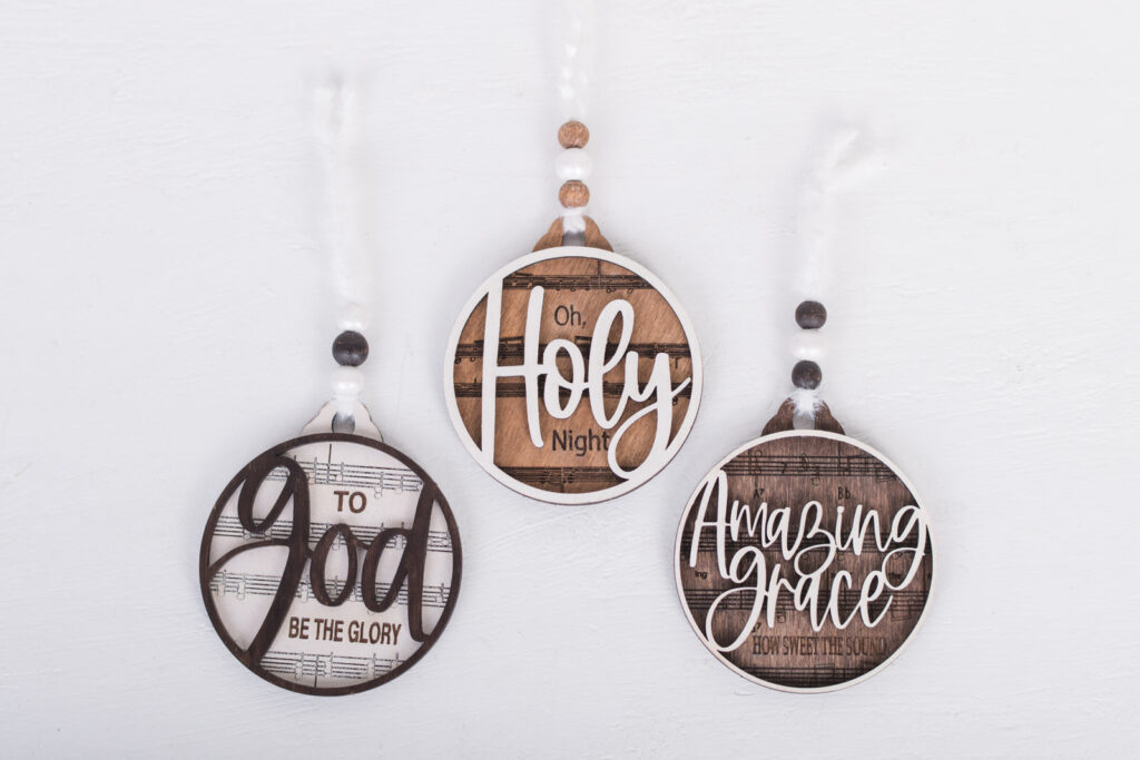 DIY Hymn Ornaments featuring ‘To God Be the Glory,’ ‘Oh Holy Night,’ and ‘Amazing Grace’ with layered wood design and sheet music background.