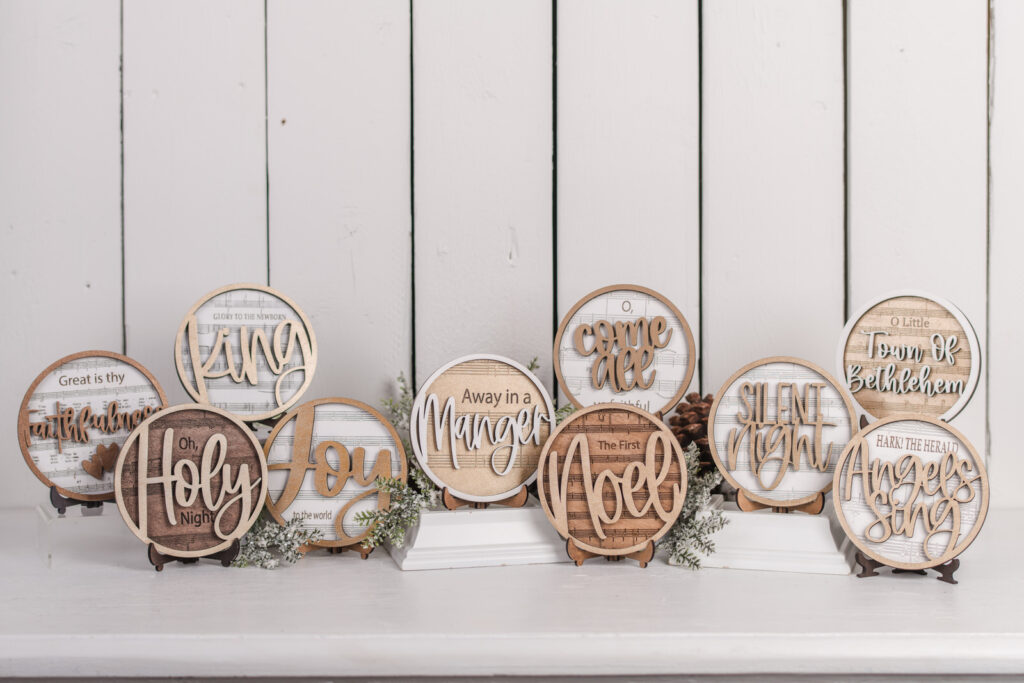 DIY Hymn Shelf Sitter Sign collection with handcrafted wood layers and music sheet backgrounds, including ‘Faithfulness,’ ‘King,’ ‘Oh Holy Night,’ ‘Joy,’ ‘Noel,’ and ‘Silent Night.’