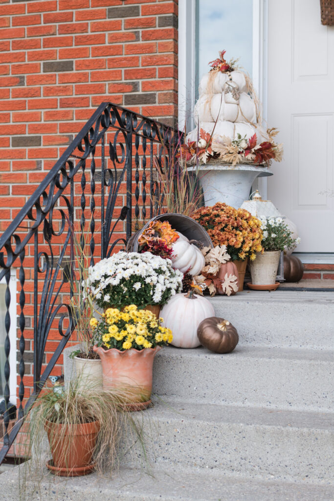 Layered pots and planters with white and rust-colored mums, pumpkins, and natural accents on front porch steps.