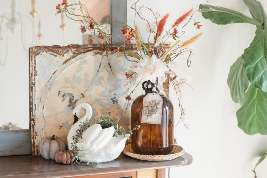 Vintage tin tile with a swan planter, and Amber glass jug with fall florals in some DIY pumpkin, sitting on top of the China cabinet in front of the Thrifted mirror for fall decor 