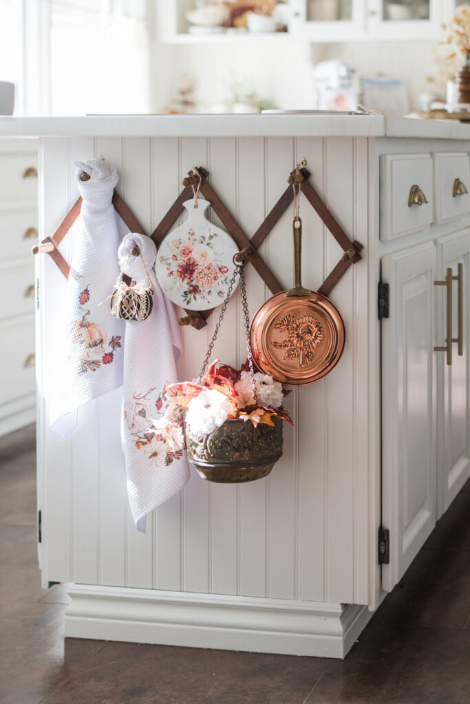 Vintage accordion hanger on the end of a kitchen island, decorated for fall with tea towels, a copper pan, a trivet, and a brass plant pot with fall florals 