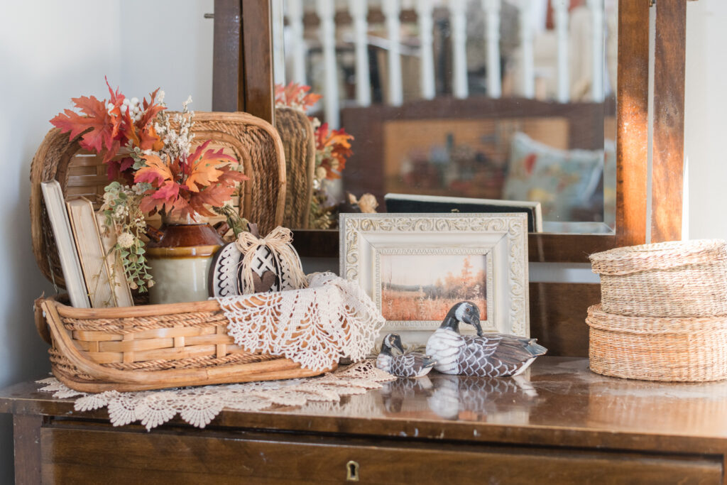 Close-up of an antique vanity styled with orange and cream leaves, a small crock, textured pumpkins in a old basket with vintage books, and two carved wooden ducks.