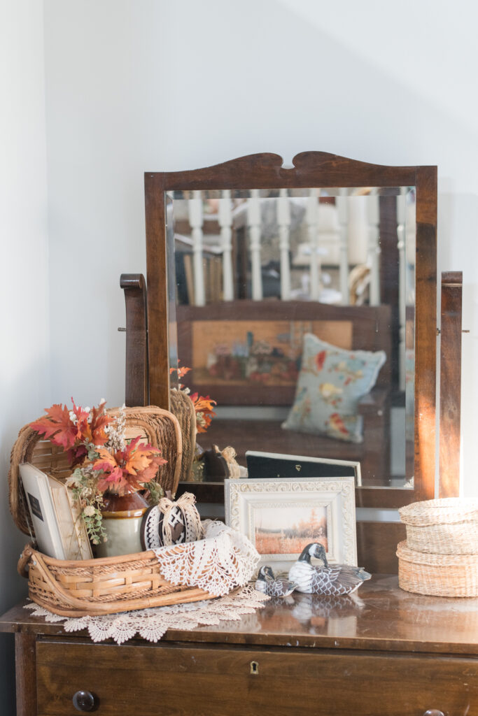 Vintage wooden dresser with a mirror, topped with a basket of books, fall florals, doily, and small pumpkins in neutral tones.