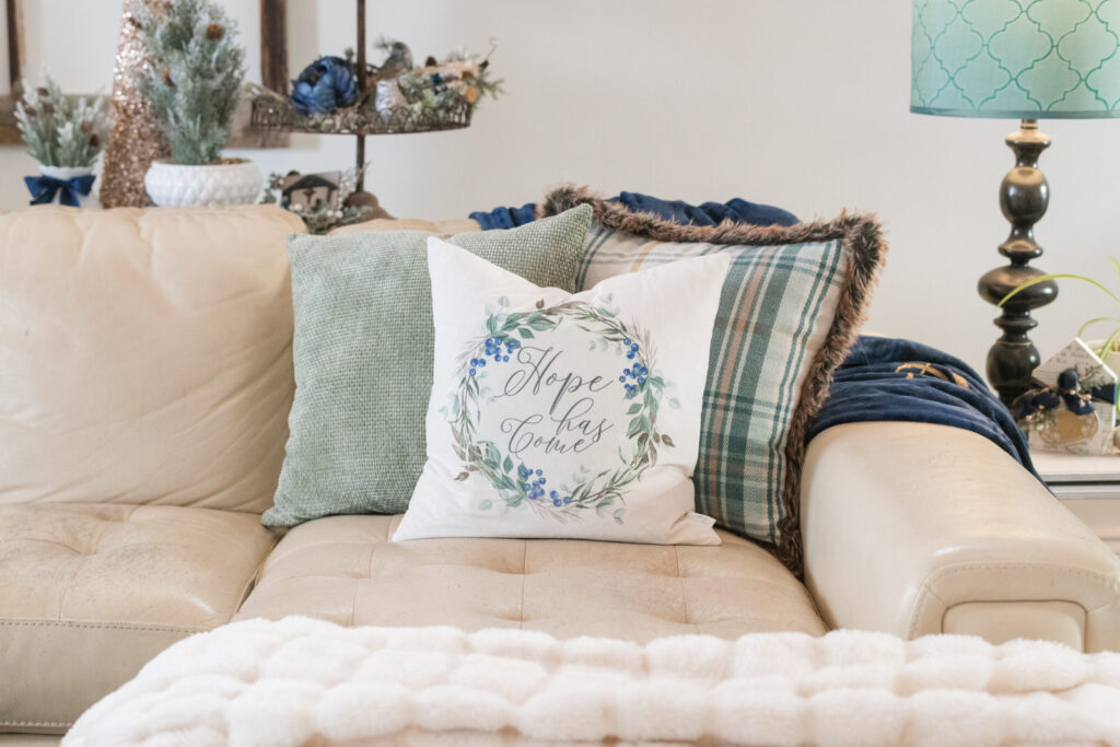 Christmas pillow arrangement on a beige sofa featuring “Hope Has Come” in blue and green with cozy plaid accents.