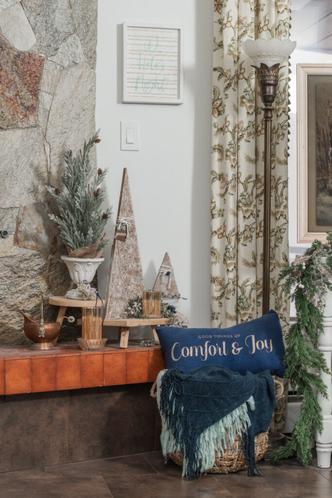 Christmas decor with a fireplace, textured trees, a basket filled with navy and turquoise blankets, and a navy pillow that says good tidings of comfort and joy styled with an antique lamp