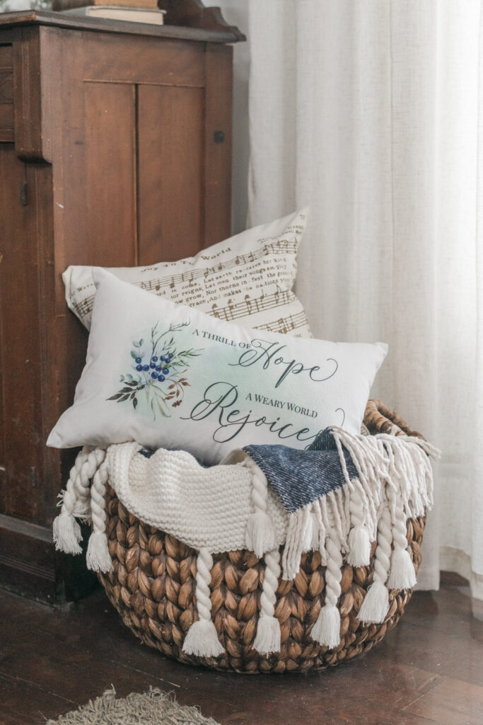 Woven basket with Christmas pillows and knit throw styled beside a wooden cabinet for a cozy winter look.
