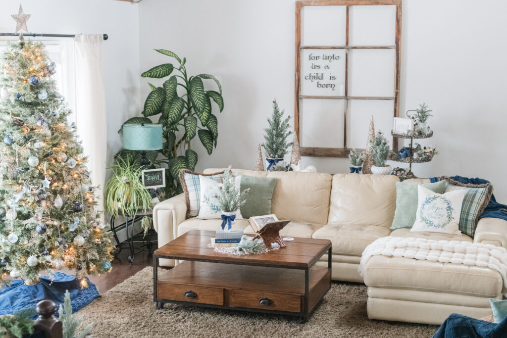 Cozy Christmas living room with neutral sofa, mini trees, and layered green and plaid pillows featuring faith-inspired Christmas designs.