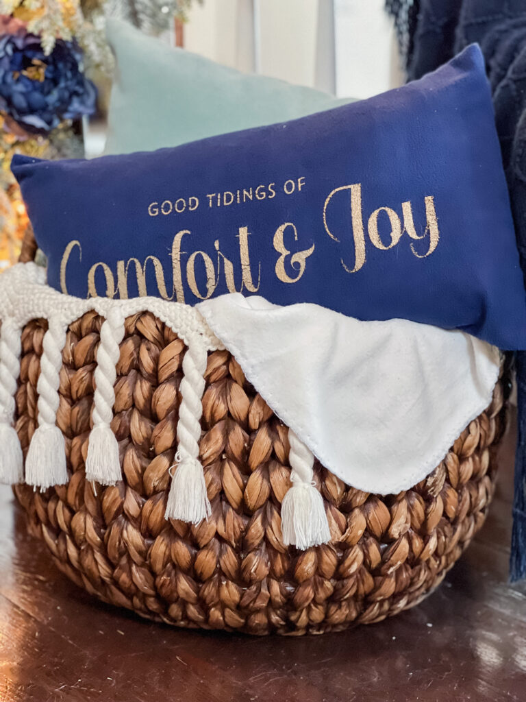 A woven basket with some cream throws inside of it is a soft, turquoise, plain pillow, and a good tidings of comfort and joy, navy velvet pillow