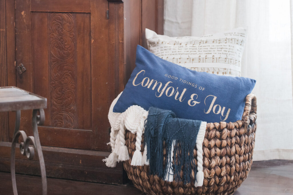 A white pillow with joy to the world Christmas music, and a blue velvet pillow that says good tidings of comfort and joy sitting in a basket with a white and navy blanket blankets draped over