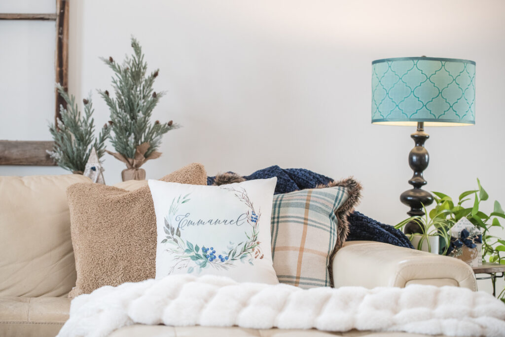 Cream, leather sofa, styled with textured pillows and Emmanuel pillow cover with a turquoise and brown lamp and faux trees in the background with a cream throw