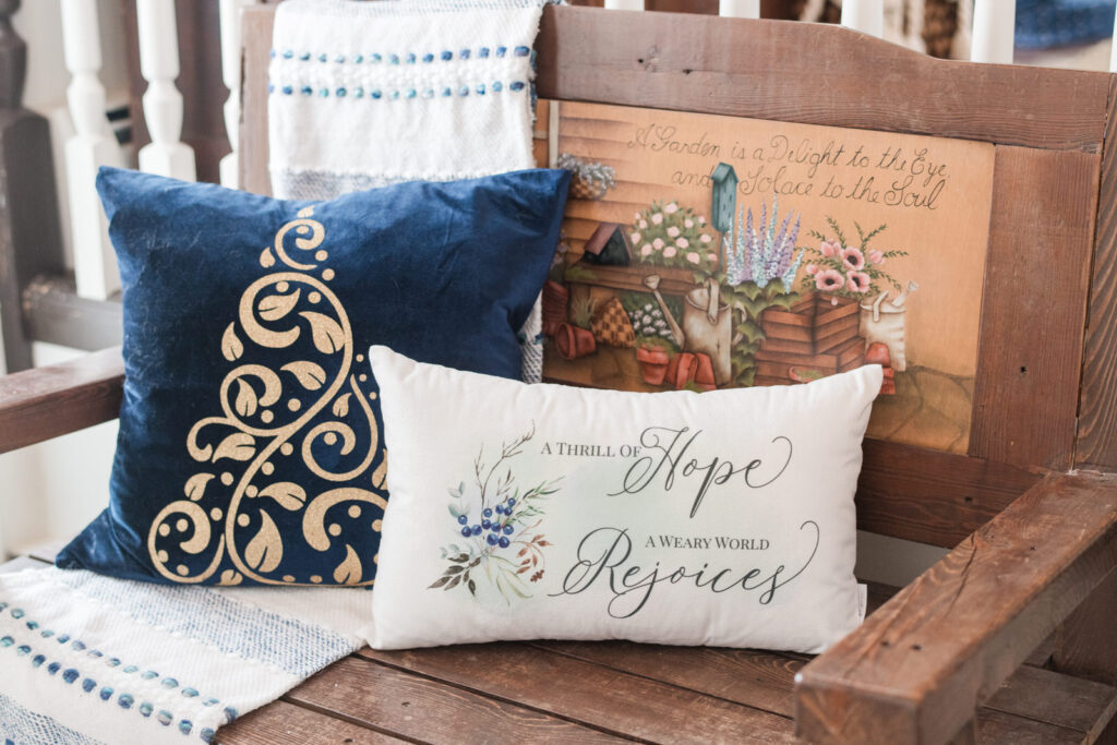 Blue velvet pillow with gold glitter Christmas tree, and a white velvet pillow that says a thrill of hope, a weary world rejoices sitting on a front entrance bench