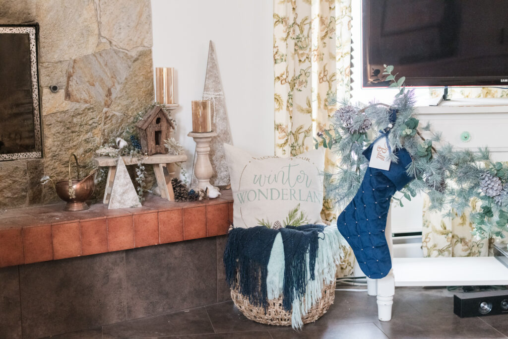 Winter wonderland pillow in a basket with navy and turquoise throws sitting beside a decorated fireplace and TV hutch with navy stockings and Garland