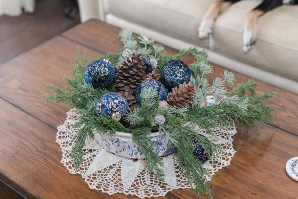 Coffee table style for Christmas with a white and blue bowl sitting on top of a vintage doily with greenery, pinecones, and blue and gold velvet ornaments 