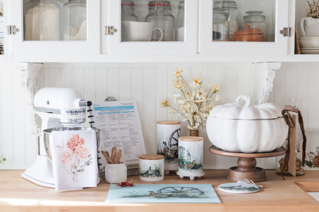 Heritage pheasant canisters beside a white KitchenAid mixer with a hydrangea tea towel, a white pumpkin tureen on a wooden pedestal, and a glass cutting board