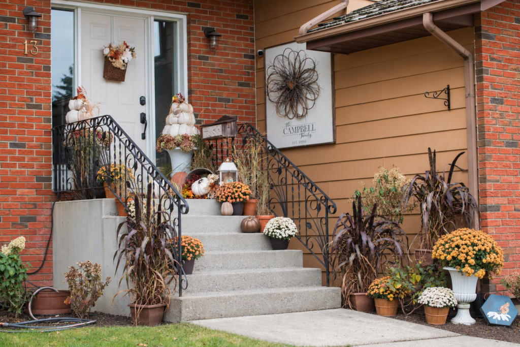 Autumn front step decor with drop-cloth pumpkin topiaries, staggered orange and cream mums, tall purple millet, and potted grasses on a brick entrance