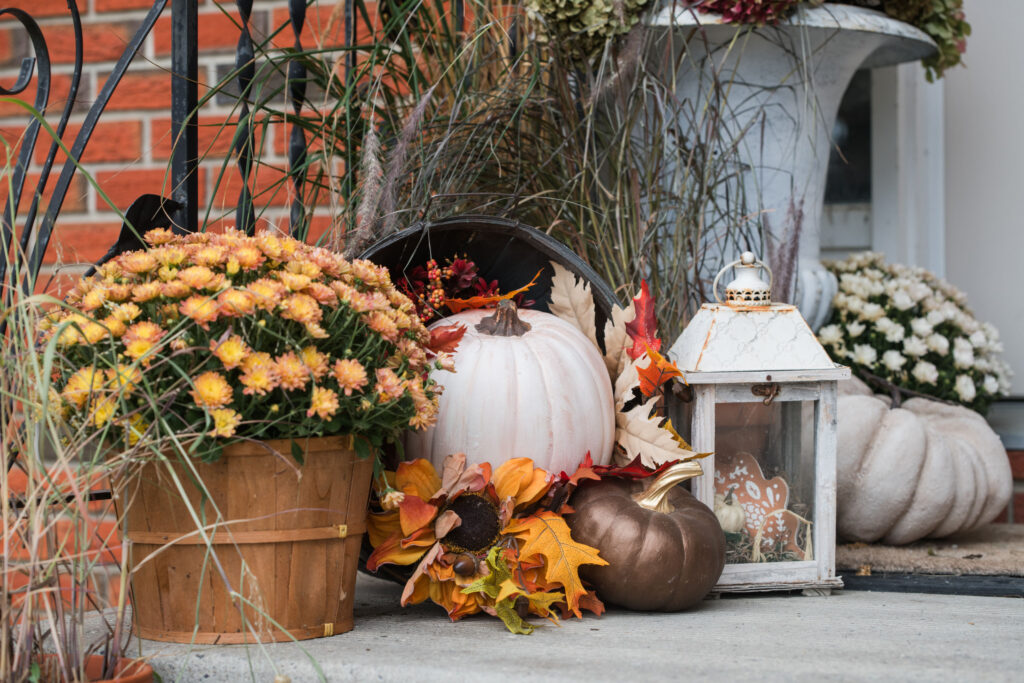 Tipped apple crate spilling faux pumpkins and autumn leaves beside a white lantern and mums on a front step