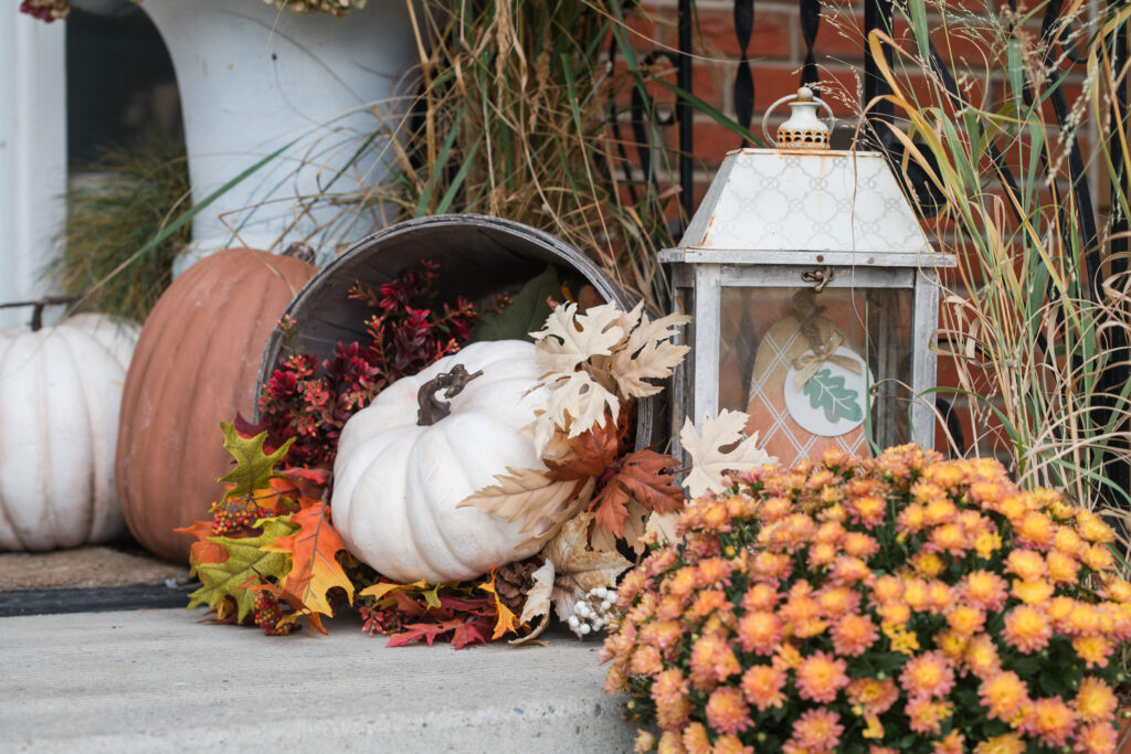 Tipped apple crate spilling faux pumpkins and autumn leaves beside a white lantern and mums on a front step