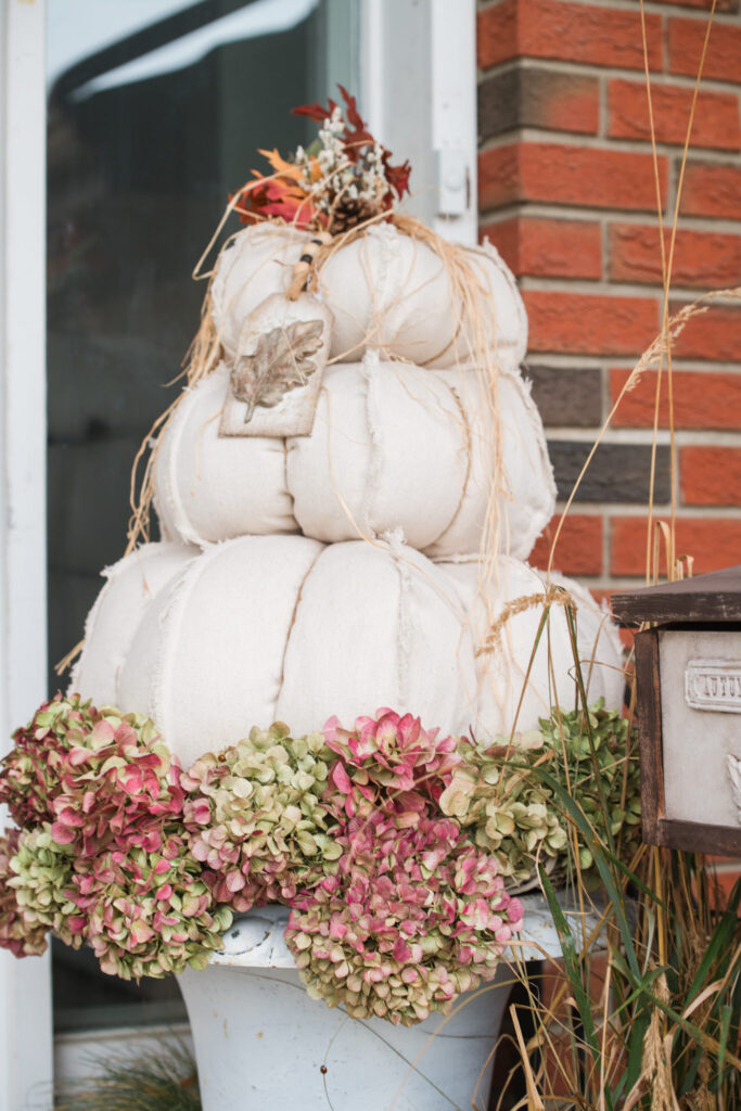 drop-cloth pumpkin topiaries on a front step, one on a white urn with dried hydrangea blooms, beside a brick wall