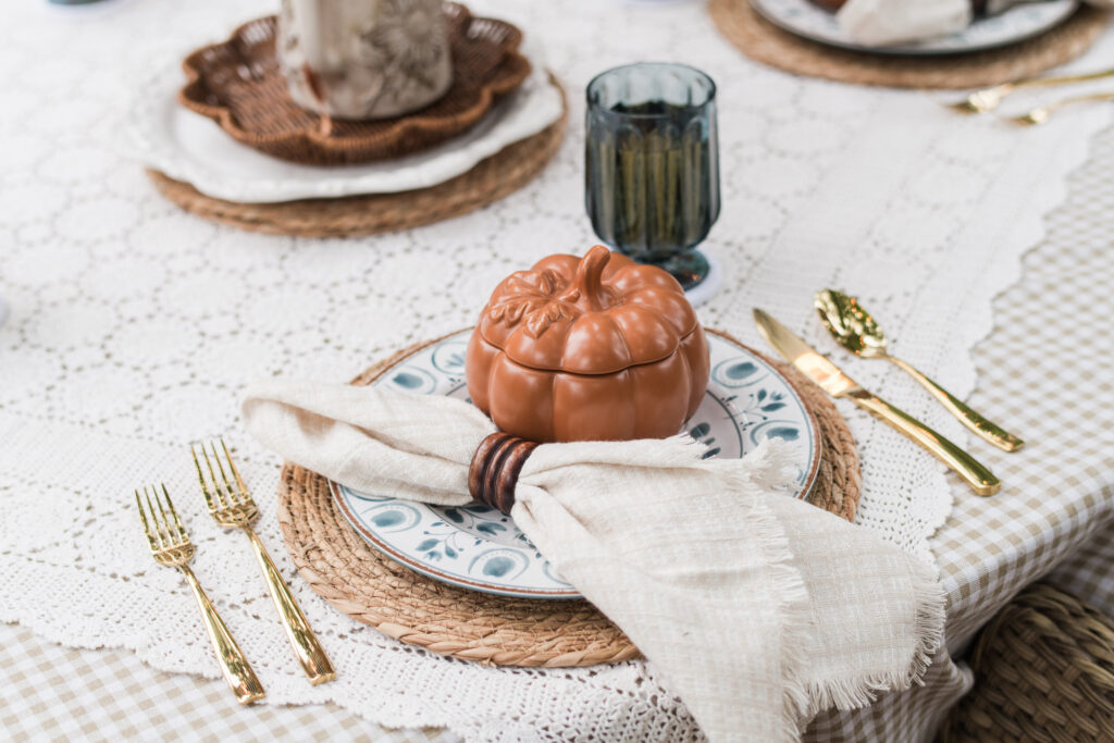 Close-up of an outdoor place setting with a blue patterned dish, gold flatware, a smoky blue glass, and a soft orange pumpkin bowl