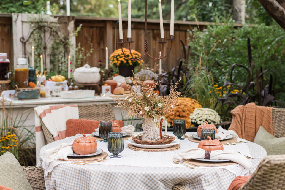 Round outdoor table under an apple tree set with layered cream and checked tablecloths, a crocheted cloth, woven placemats, blue melamine dishes, and soft orange pumpkin bowls