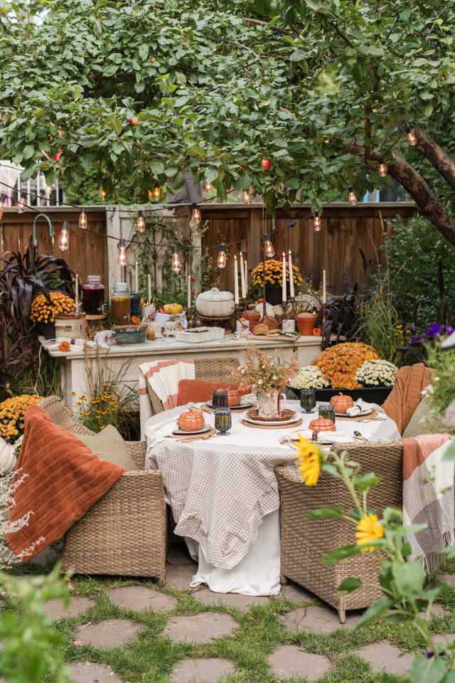 Round outdoor table under an apple tree set with a checked tablecloth, a crocheted cloth, woven placemats, blue dishes, and soft orange pumpkin bowls
