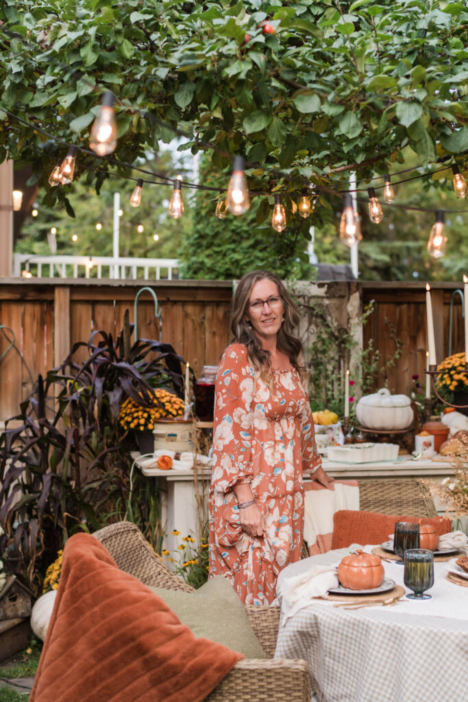 Kimberly standing under string lights beside an outdoor dinner table set for an autumn harvest gathering