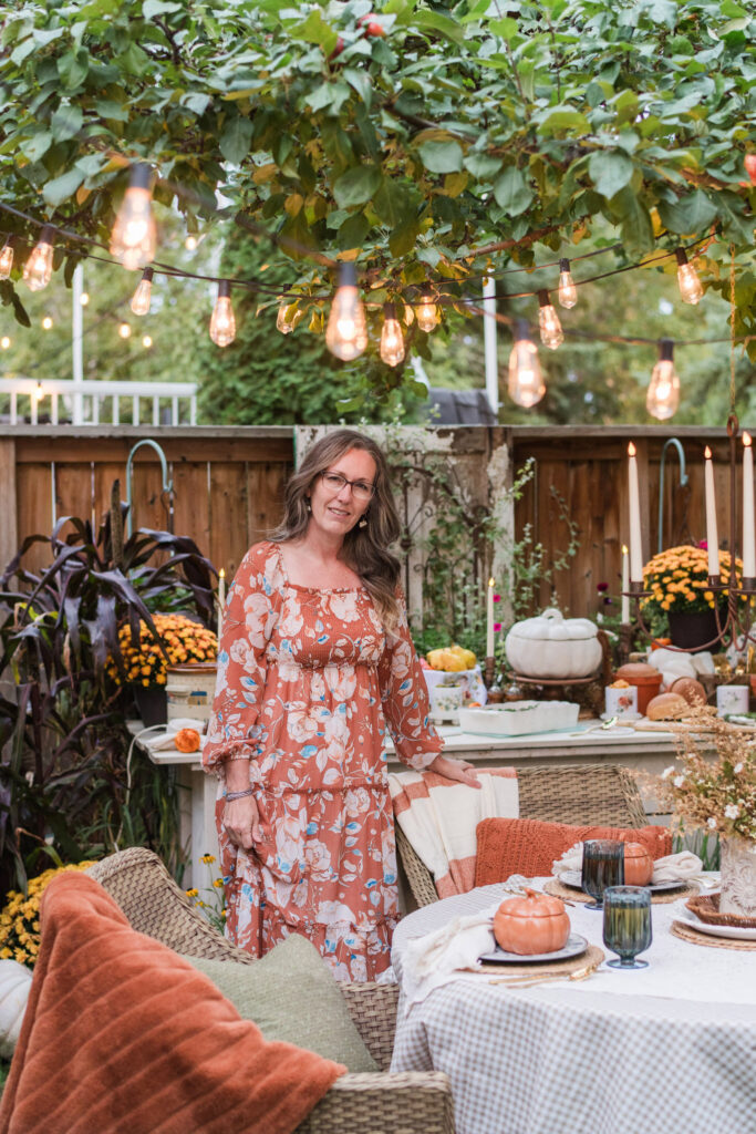 Kimberly standing under string lights beside an outdoor dinner table, welcoming you to an autumn home tour
