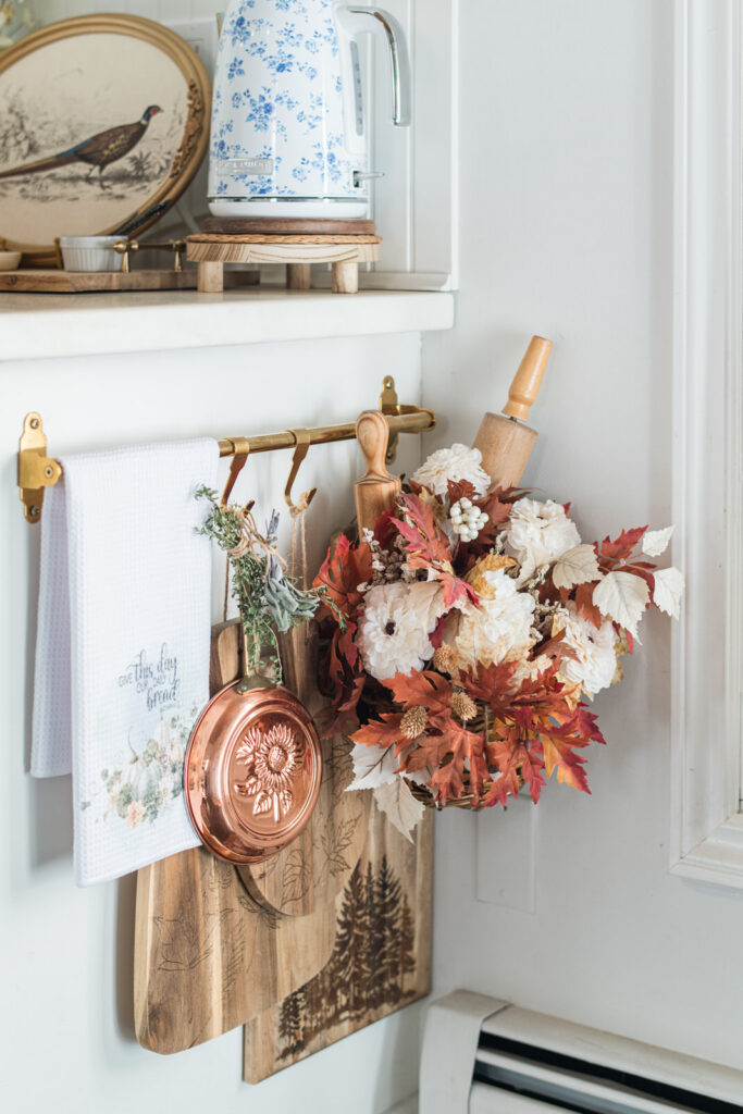 The end of the counter decorated with a hanging basket full of rolling pins, fall florals, layered cutting boards, a copper pan and a tea towel