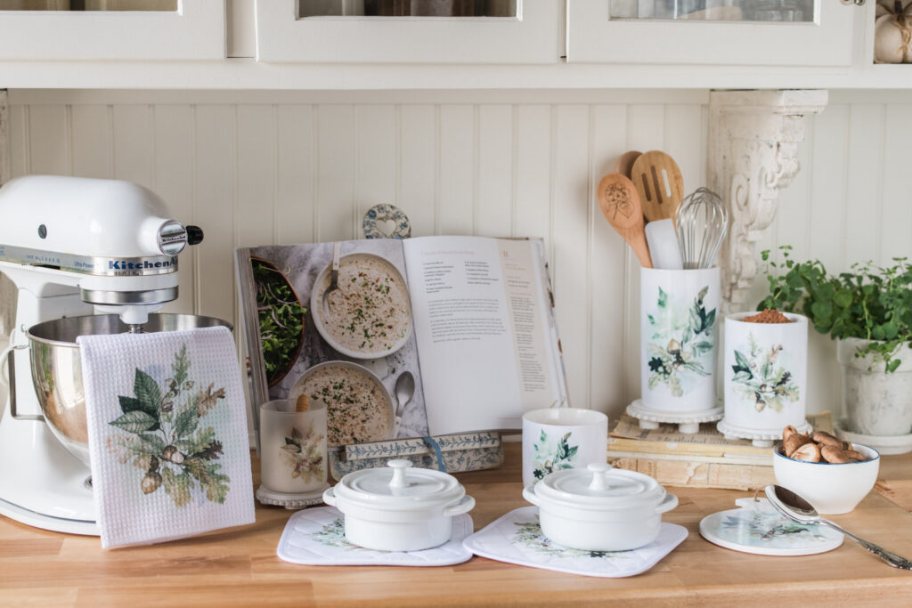 Two white mini cocottes on Woodland potholders beside a Woodland tea towel, canisters, and an open cookbook