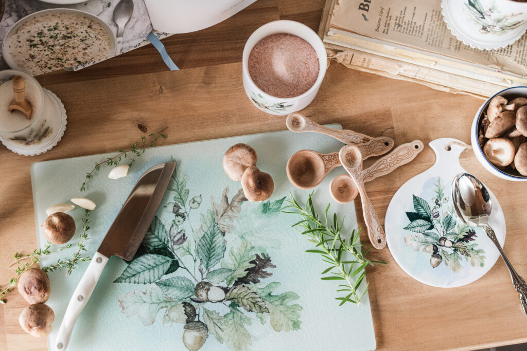Overhead view of a Woodland glass cutting board with a knife, fresh herbs, wooden measuring spoons, and a Thankful trivet beside old cookbooks